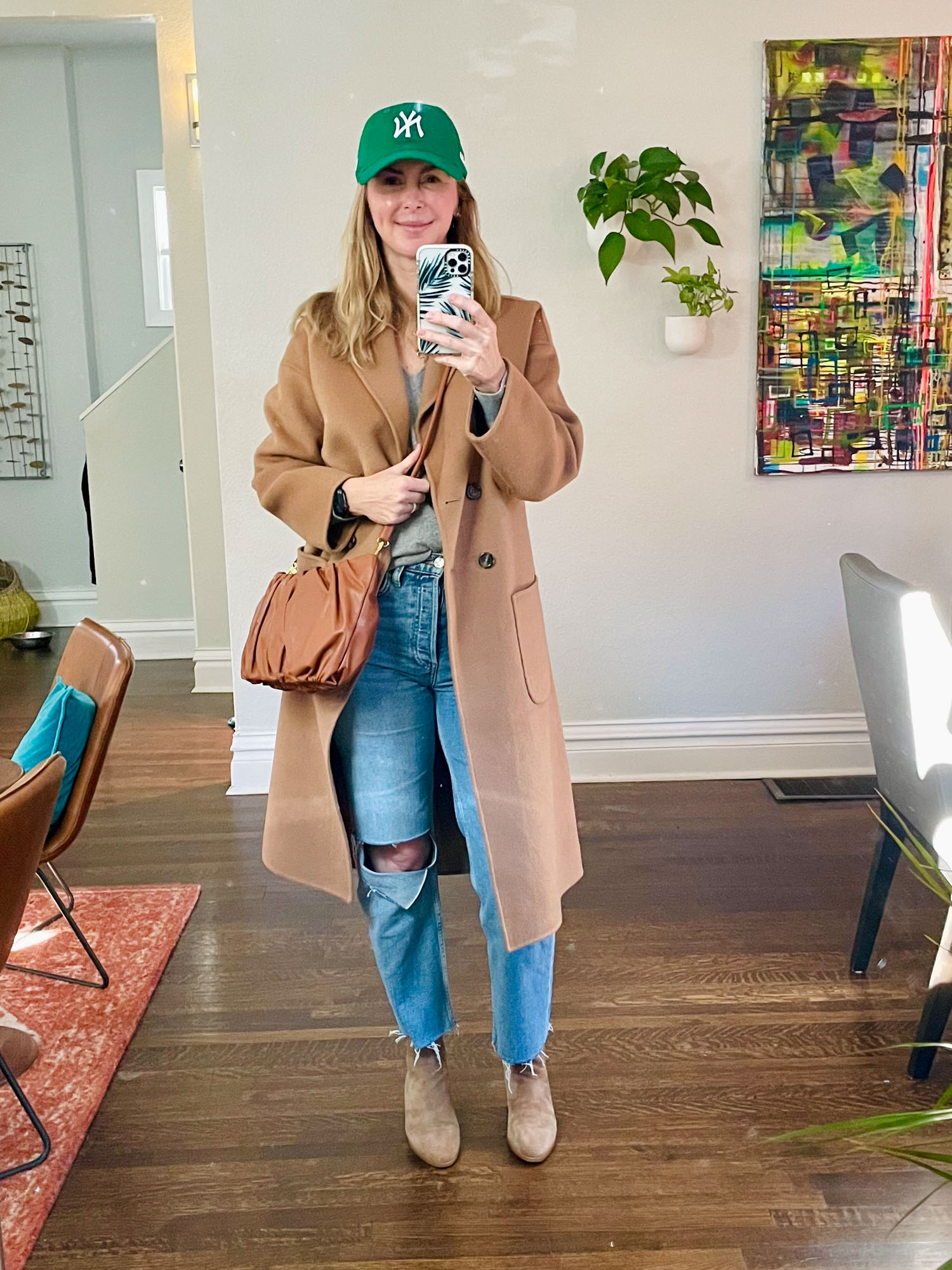 Wearing the Anine Bing camel coat casually with distressed jeans, a gray swater and a green NY Yankees baseball cap.