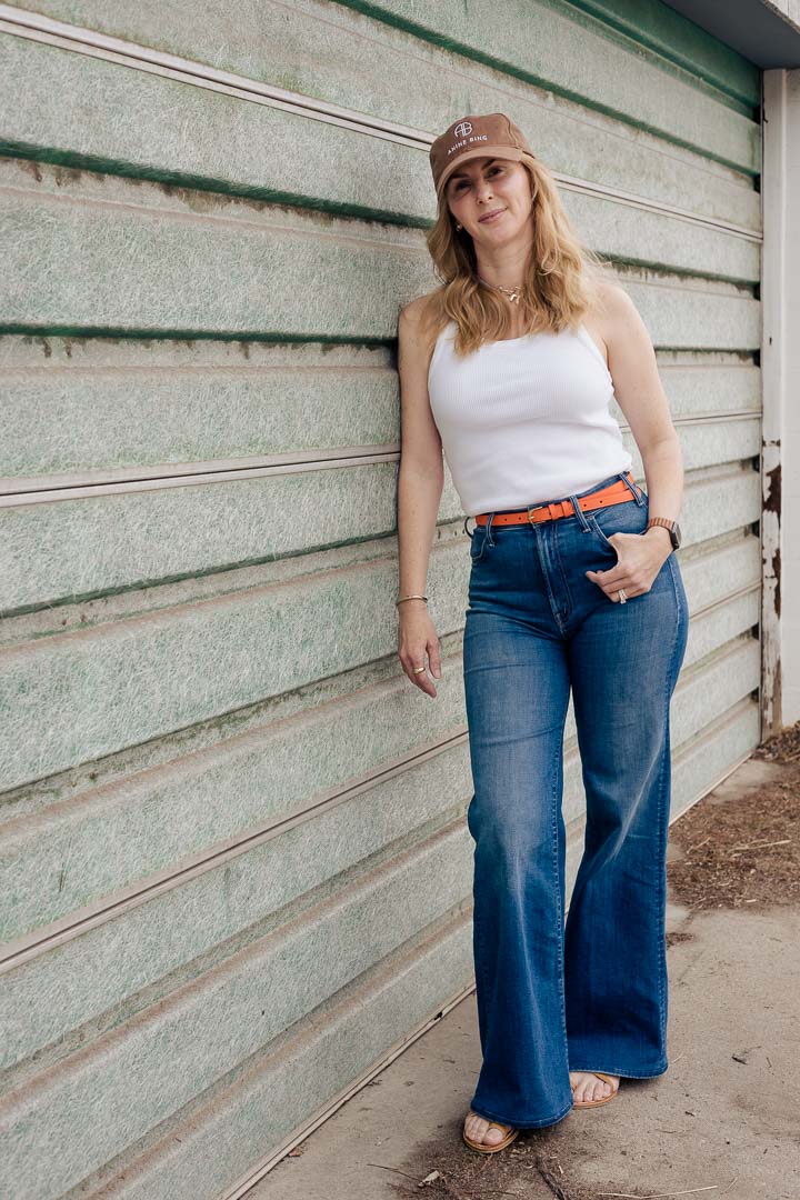 Wearing the Anine Bing Jeremy baseball cap in dark camel with a white Agolde tank top, orange belt by Frame, and MOTHER Hustler roller jeans.