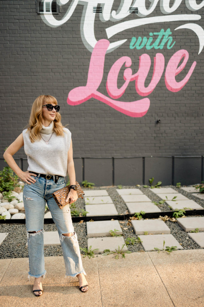 Standing in front of a Love mural wearing a gray, sleeveless cashmere Free People sweater with distressed Moussy Odessa jeans, a black belt, heels and a leopard bag.