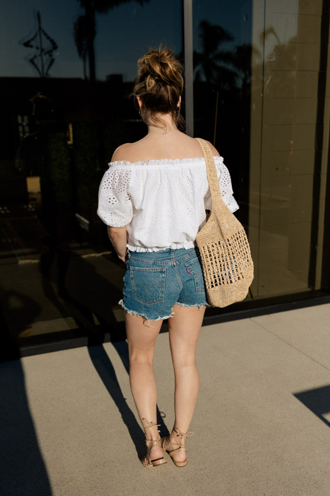 In Solana Beach, CA, wearing spring outfit of white top and denim shorts and raffia bag.