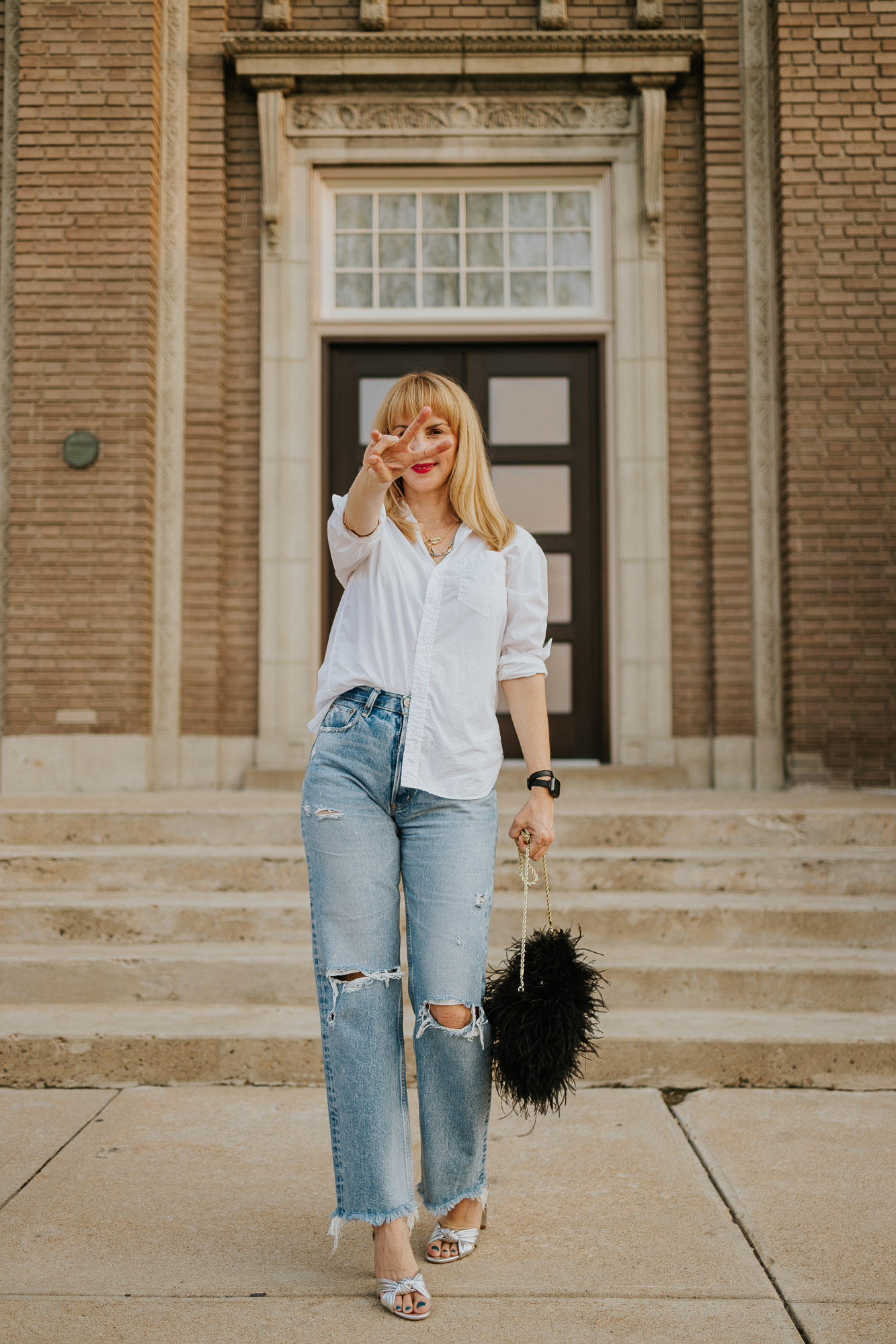 Wearing a white button down Frank & Eileen shirt with Moussy jeans, Veronica Beard silver heels and a black feather bag making a peace sign.